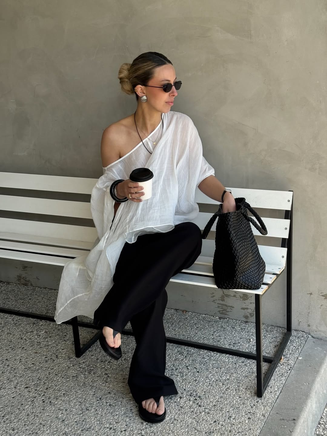 Woman in sheer white linen one-shoulder tunic and black wide-leg flare trousers with woven leather tote, what to wear in NYC in summer