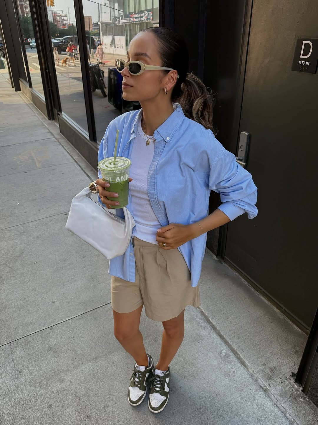 Woman in oversized periwinkle oxford shirt and khaki Bermuda shorts with Nike Dunks and white shoulder bag, casual NYC summer outfit on a downtown street