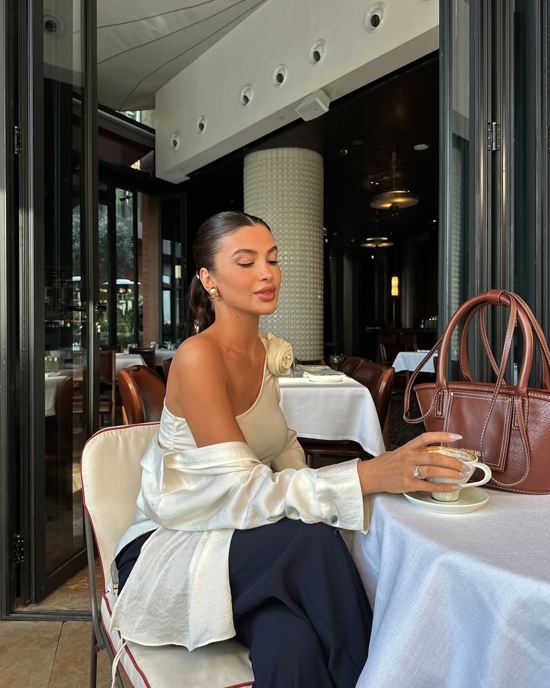 Woman in ivory one-shoulder satin top with rose appliqué and navy trousers at a restaurant table, elegant NYC summer look