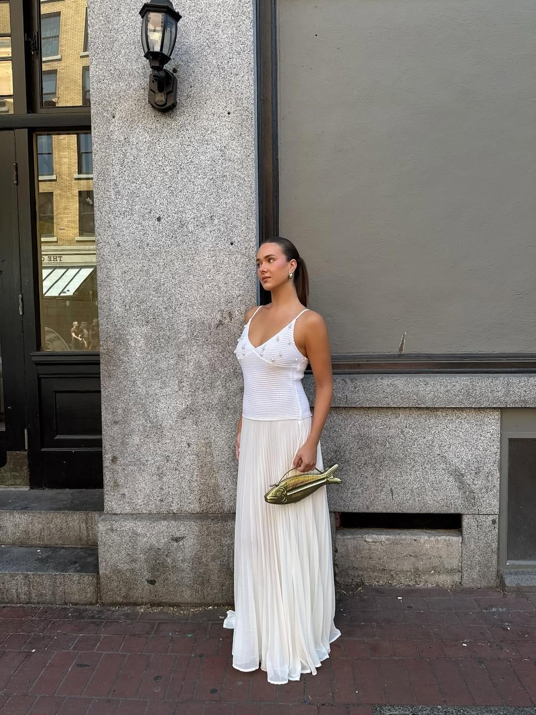 Woman in pearl-embellished white mesh cami and white accordion-pleated maxi skirt holding gold fish clutch outside a NYC building, summer outfit for New York