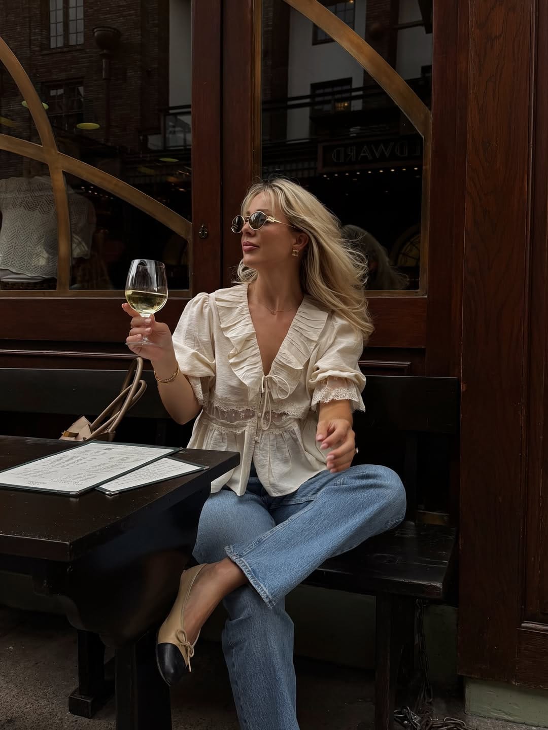  Blonde woman in cream ruffle lace blouse and light blue straight jeans with two-tone ballet flats at an outdoor bistro, summer outfit for New York