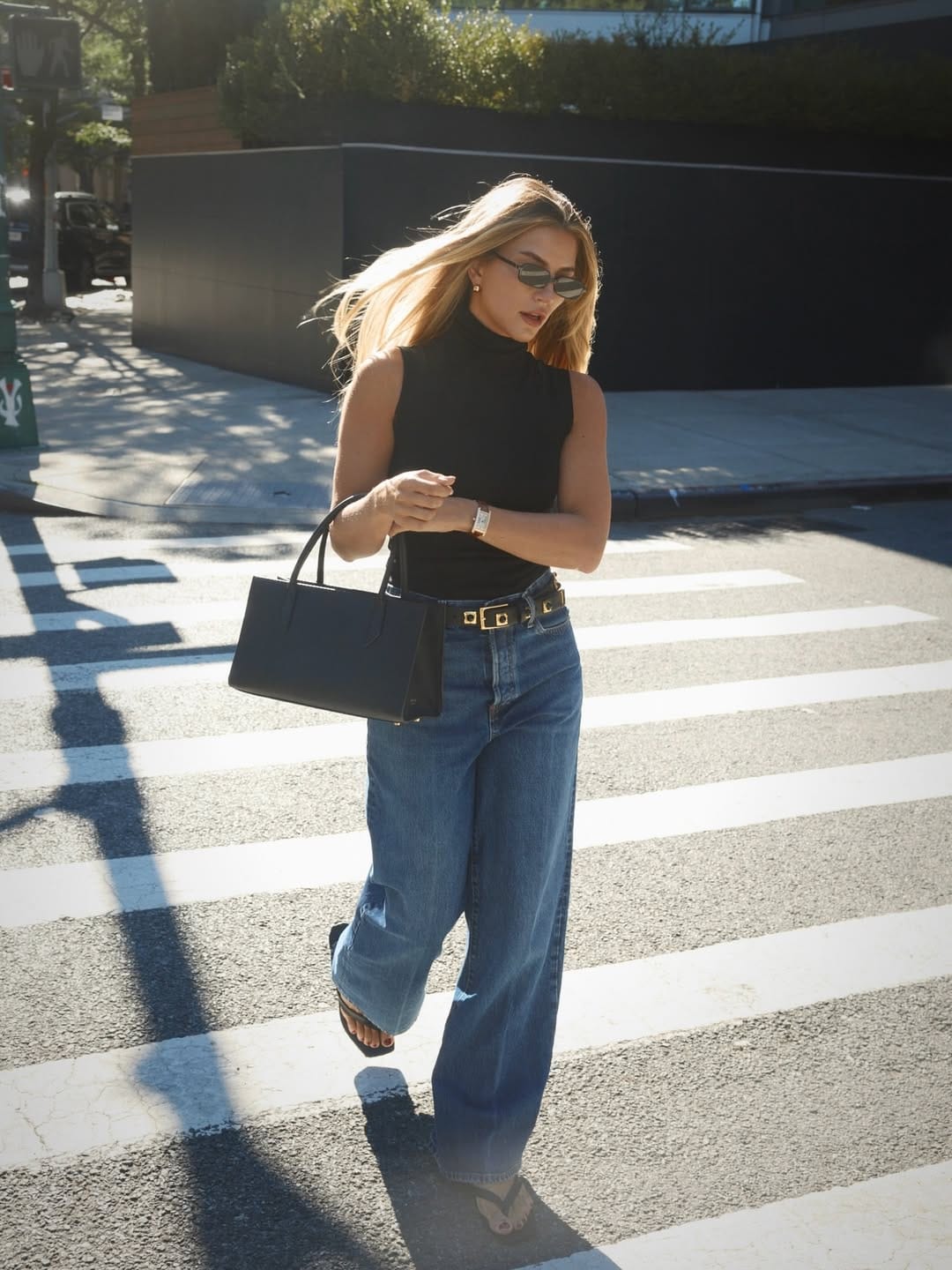 Blonde woman in black mock-neck sleeveless top and wide-leg blue jeans with gold square-buckle belt crossing a sunlit New York street