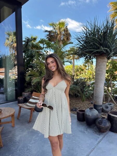 Woman in a sage and white striped sweetheart mini dress holding sunglasses on a palm-lined patio