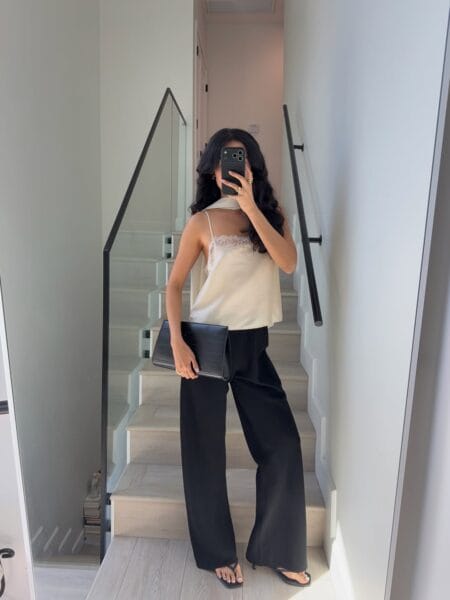 Mirror selfie of a woman in a champagne silk cami and black wide-leg trousers with a clutch