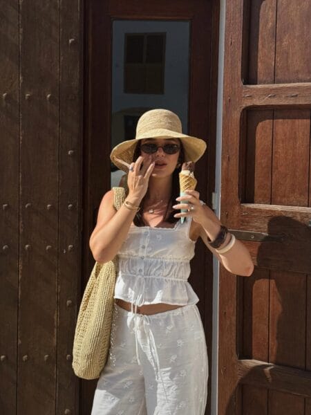 Woman in a white broderie peplum top and trousers with a wide straw hat and raffia bag