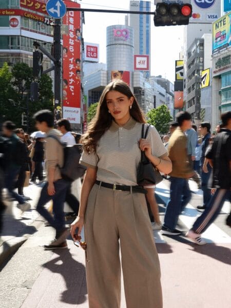 Woman in a sage striped polo and taupe wide-leg trousers crossing a busy city street