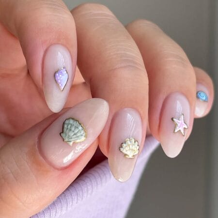 Sheer nude almond nails with gold-outlined coastal charm stickers including shells, starfish and a spiral motif — euro summer nail idea