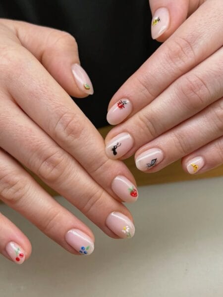 Short oval cream nails with tiny micro-painted garden illustrations including a bee, ladybird, ant, butterfly, strawberry and cherries