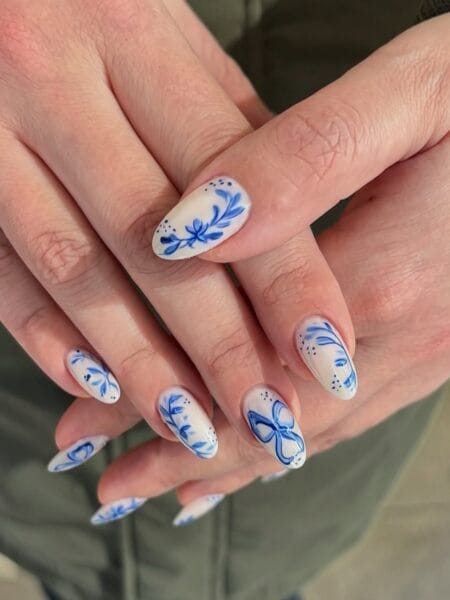 Frosted white almond nails with cobalt blue hand-painted bows, botanical branches and butterfly motifs in Delft porcelain style