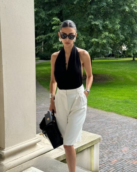 Black deep-V halter bodysuit and white tailored Bermuda shorts with silver cuff bracelets and structured bag — trendy summer looks