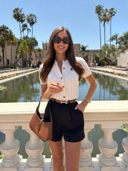 White cable-knit short-sleeve cardigan and black tailored cuffed shorts with tan belt and cognac shoulder bag — june outfits 2026