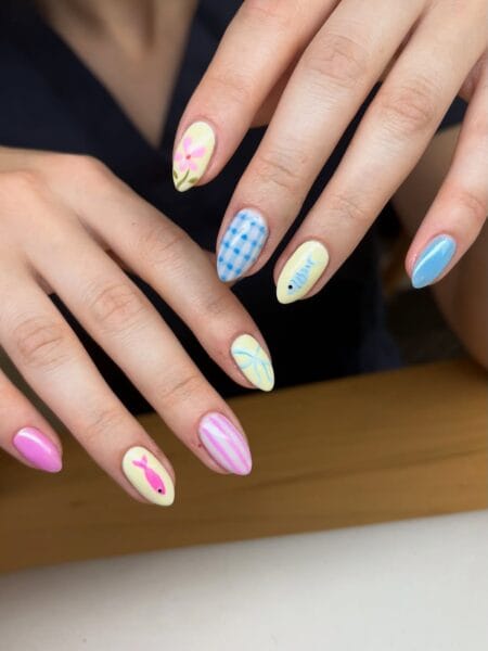 Medium almond nails in butter yellow with pink fish silhouette, blue outline fish, blue gingham check and pink stripe designs — butter yellow fish and gingham pastel nails 2026