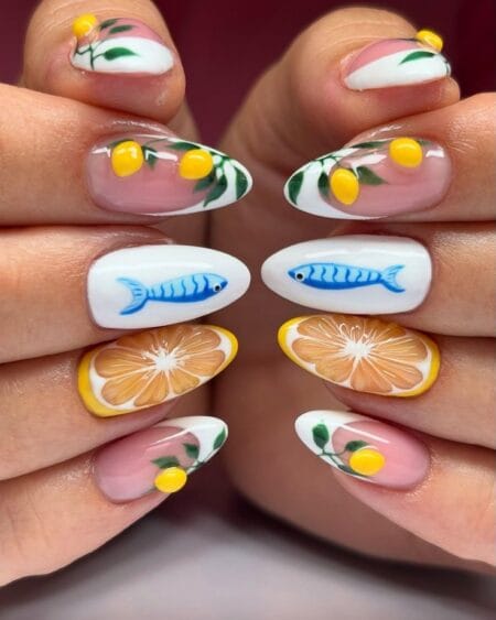 Long almond nails with sheer French tips painted with yellow lemon branches, solid white nails with blue sardine illustration and orange citrus slice nail — blue sardine and lemon botanical nails 2026