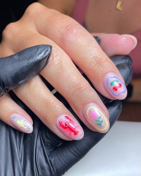 Short round sheer nude nails with hand-painted yellow fish, red lobster, pink floral cameo and cherry designs — coastal curio fish nails 2026