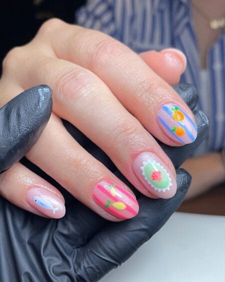 Short oval nails combining a tiny blue fish on nude base, pink stripe with lemons, shell stamp cameo and blue stripe with citrus — coastal fish nail art 2026