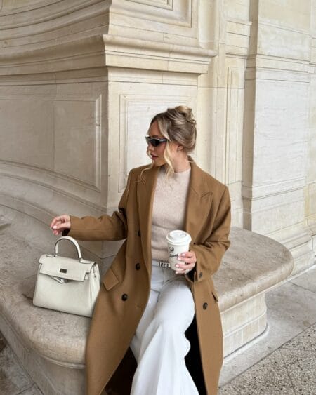 Woman in a camel wool coat, oatmeal ribbed knit and white wide-leg trousers with a cream top-handle bag