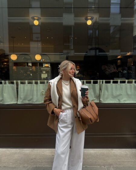 Woman in a tan suede shearling jacket with an ivory jumper and wide white trousers