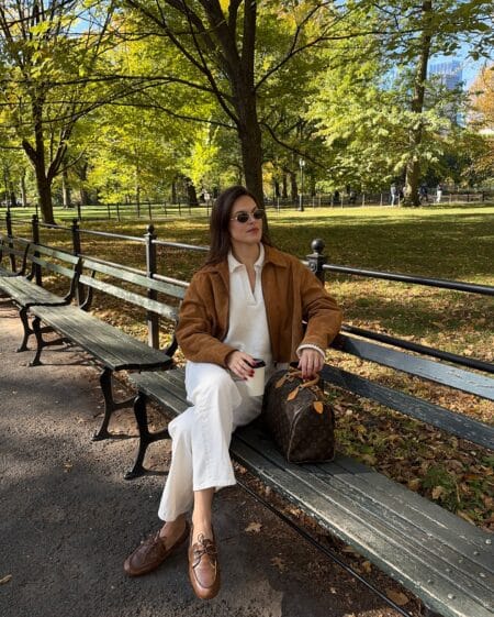Woman in a tan suede bomber with a cream polo knit, cropped white jeans and brown driving loafers