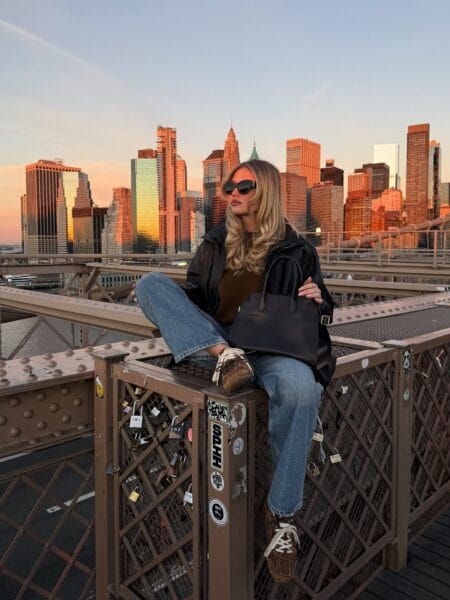 Woman in a black leather bomber with an olive knit, light wash jeans and brown suede trainers
