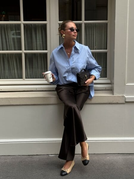 Woman in an oversized pale blue poplin shirt with chocolate satin flared trousers and cap-toe flats