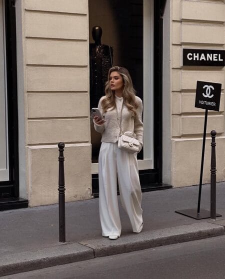Woman in a cream knit cardigan and ivory pleated trousers with a cream quilted bag and pale flats