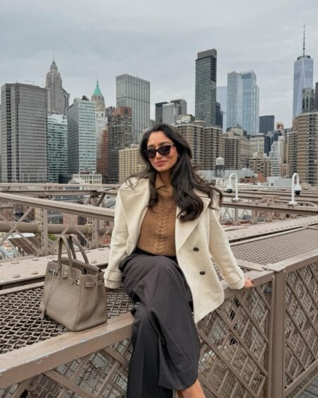 Woman in an ecru wool peacoat over a camel cable-knit jumper with charcoal wide-leg trousers on Brooklyn Bridge