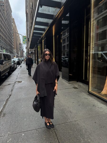 Woman in an espresso scarf-collar cape and matching brown midi dress with a woven leather pouch bag