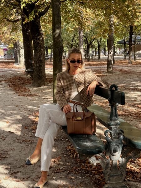Woman in brown houndstooth tweed jacket with white linen trousers and tan leather tote sitting on a Paris park bench
