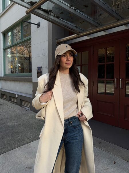 Long cream wool coat over oatmeal jumper and mid-wash jeans with a beige corduroy cap