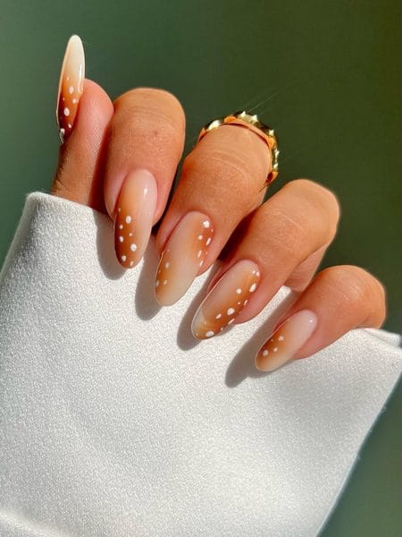 Milky beige almond nails featuring an amber aura gradient and descending white micro dots