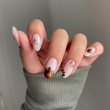 Off-white almond nails decorated with metallic bronze pumpkins, autumn flowers, and delicate starbursts